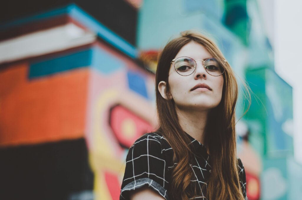 Portrait of a fashionable woman with glasses posing confidently outdoors in front of a vibrant mural.